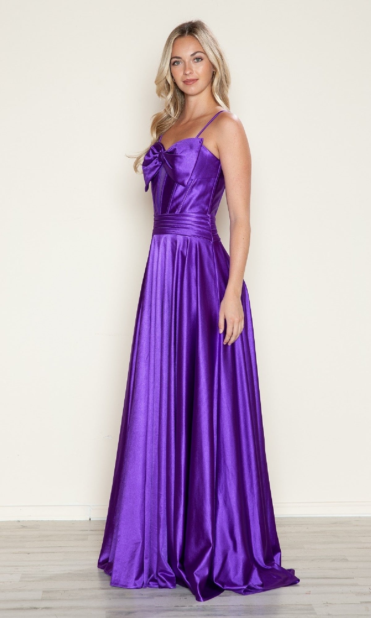 Indoors, a woman wears the Poly USA 9702 Bow-Front Long Corset Prom Dress—an elegant, floor-length purple gown featuring spaghetti straps, a shiny finish, and a bow detail on the bodice.